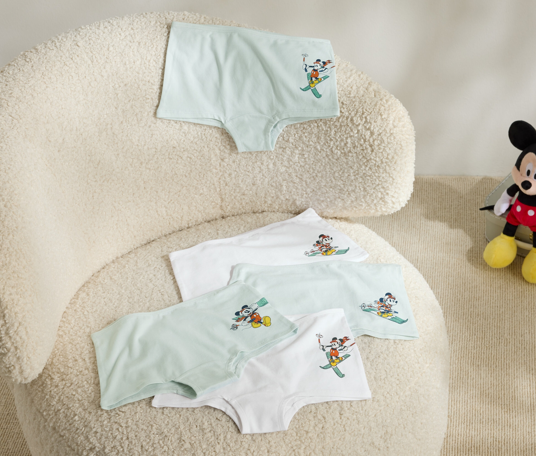 Na chlpatom kresle ležia štyri detské spodky s obrázkom Mickeyho Mousa na lyžiach a vedľa kresla sedí plyšový Mickey Mouse.