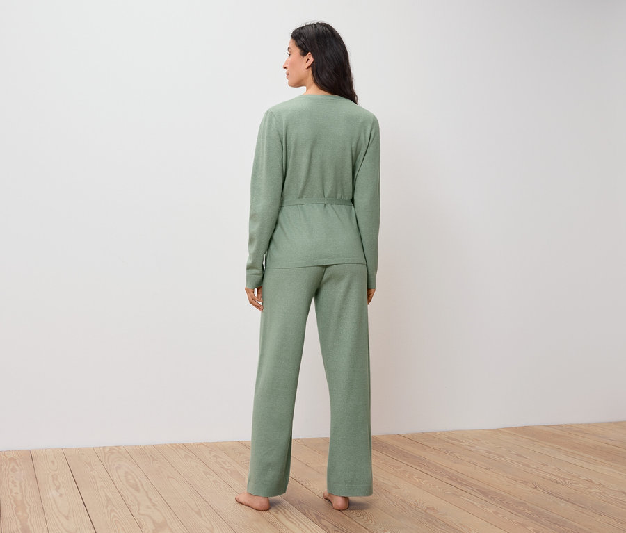 Žena v zelenom svetri a nohaviciach stojí chrbtom k divákovi na drevenej podlahe.