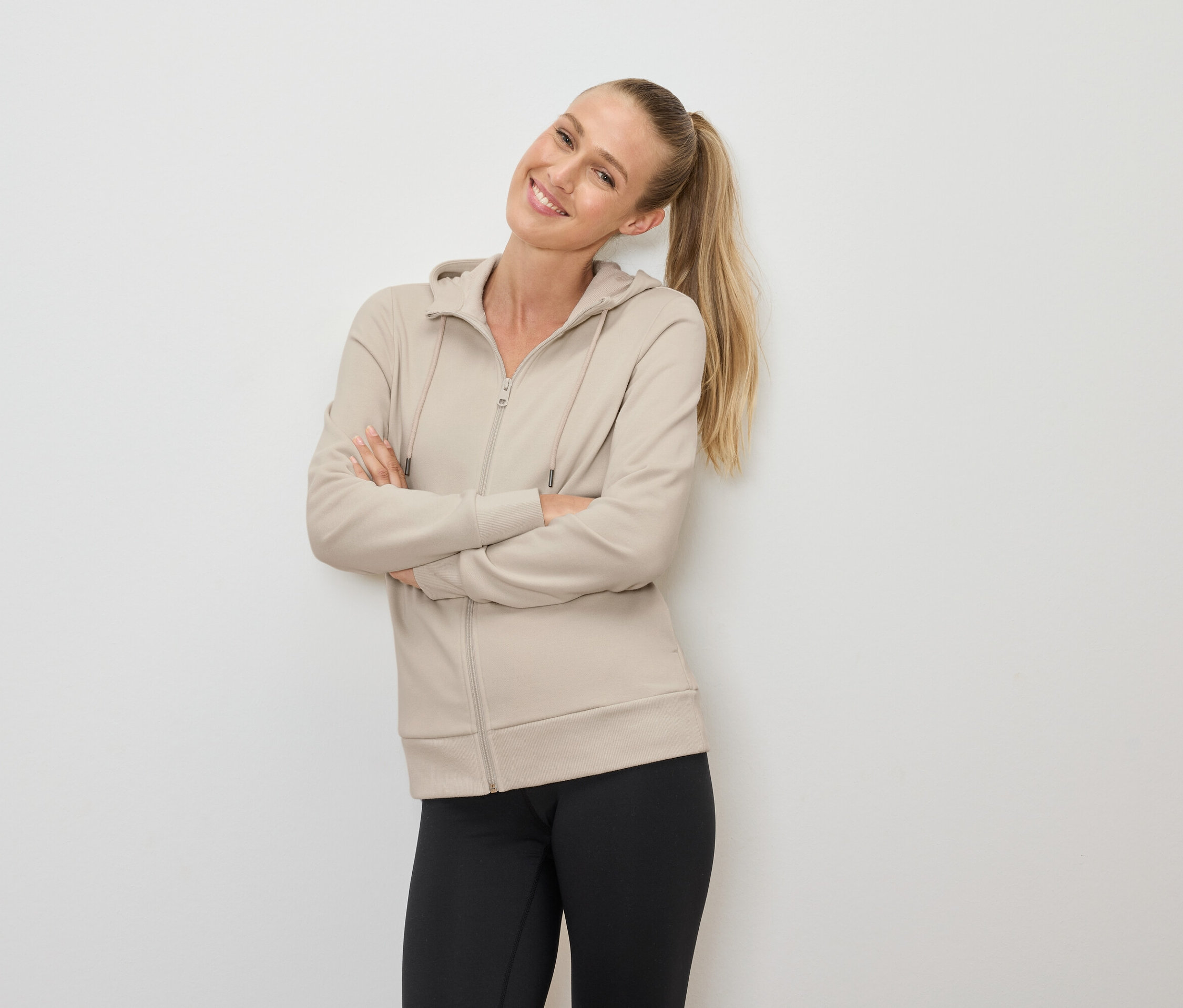 Usmievavá sa blondínka s copom pózuje so skríženými rukami, oblečená v béžovej mikine na zips a čiernych legínach.