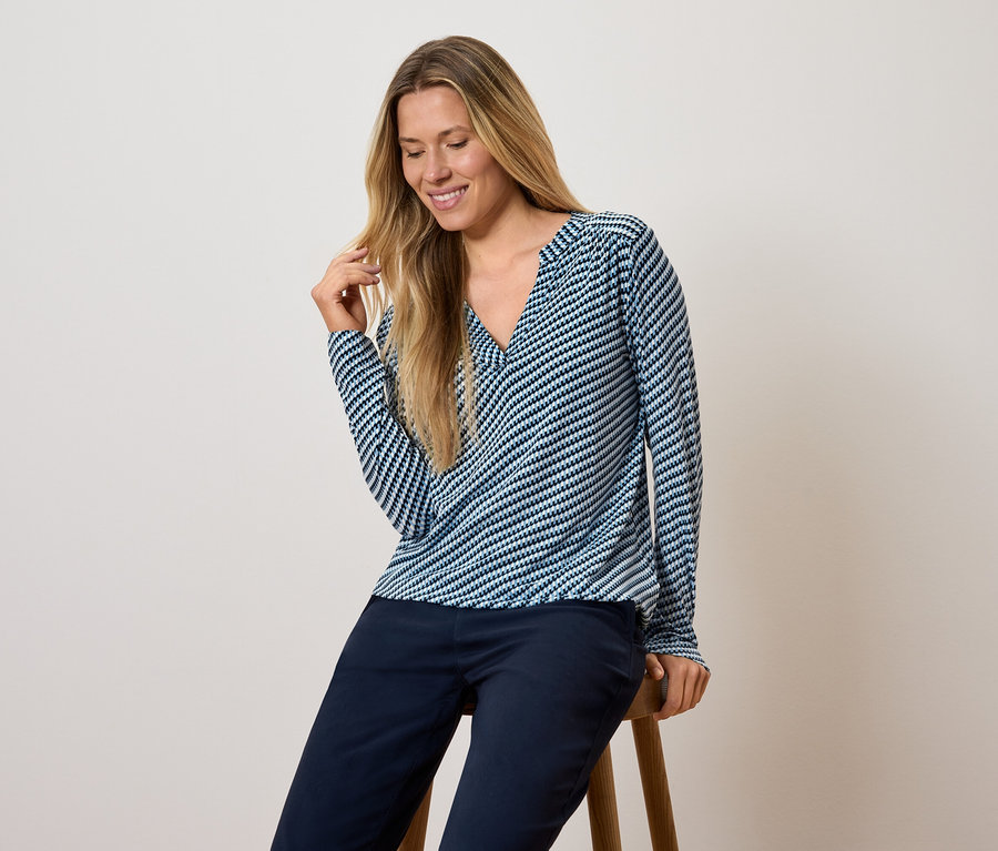 Žena sedí na stoličke, má na sebe modro-biely pruhovaný top s dlhým rukávom a tmavomodré nohavice.