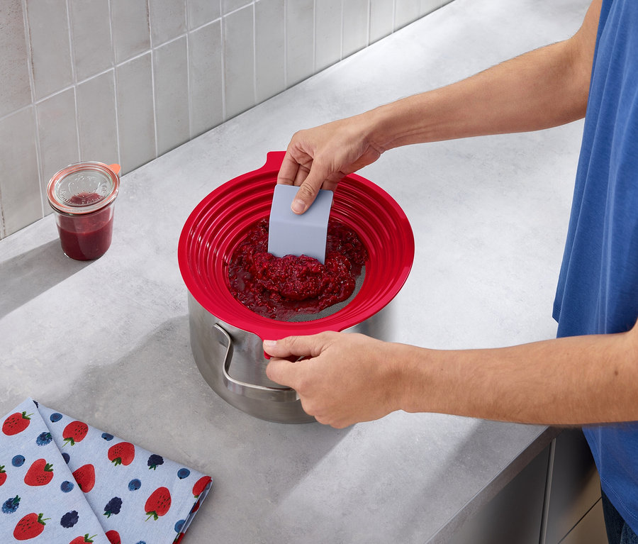 Muž pretláča malinový džem cez 2 v 1: sito na preosievanie múky a pasírovanie do hrnca pomocou sivej stierky.