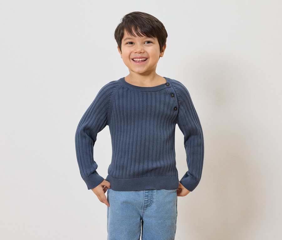 Usmievavý chlapec s hnedými vlasmi má na sebe tmavomodrý rebrovaný sveter s gombíkmi a svetlomodré džínsy.