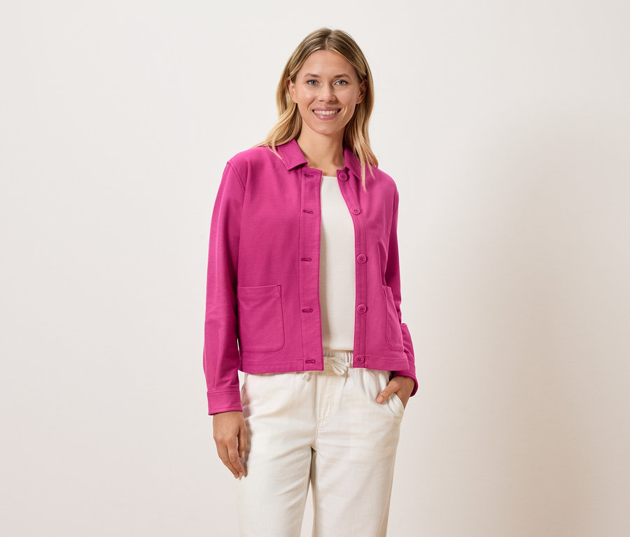 Žena má na sebe ružovú bundu cez biely top a krémové nohavice.