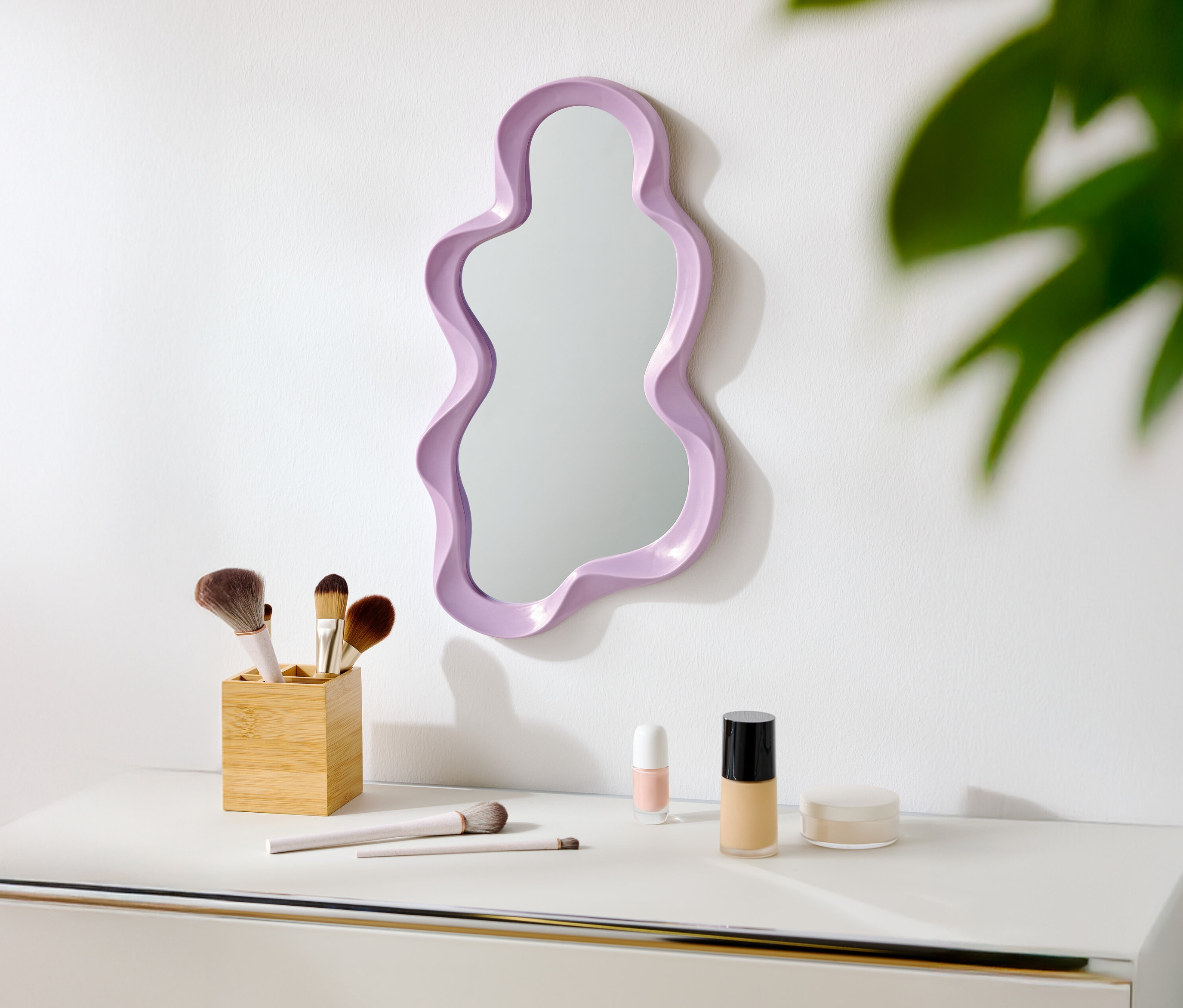 Na bielej stene visí fialové zrkadlo nepravidelného tvaru. Pod ním sú na bielom stole štetce na make-up a kozmetika.