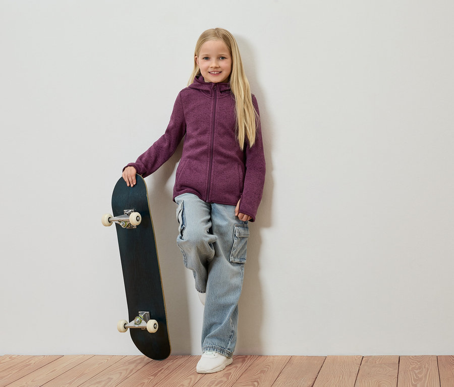 Dievča s blond vlasmi sa opiera o stenu, drží skateboard a má na sebe fialovú flísovú bundu, džínsy a biele topánky.