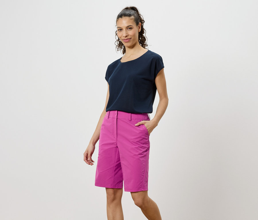 Žena v tmavom tričku a ružových šortkách stojí na bielom pozadí.