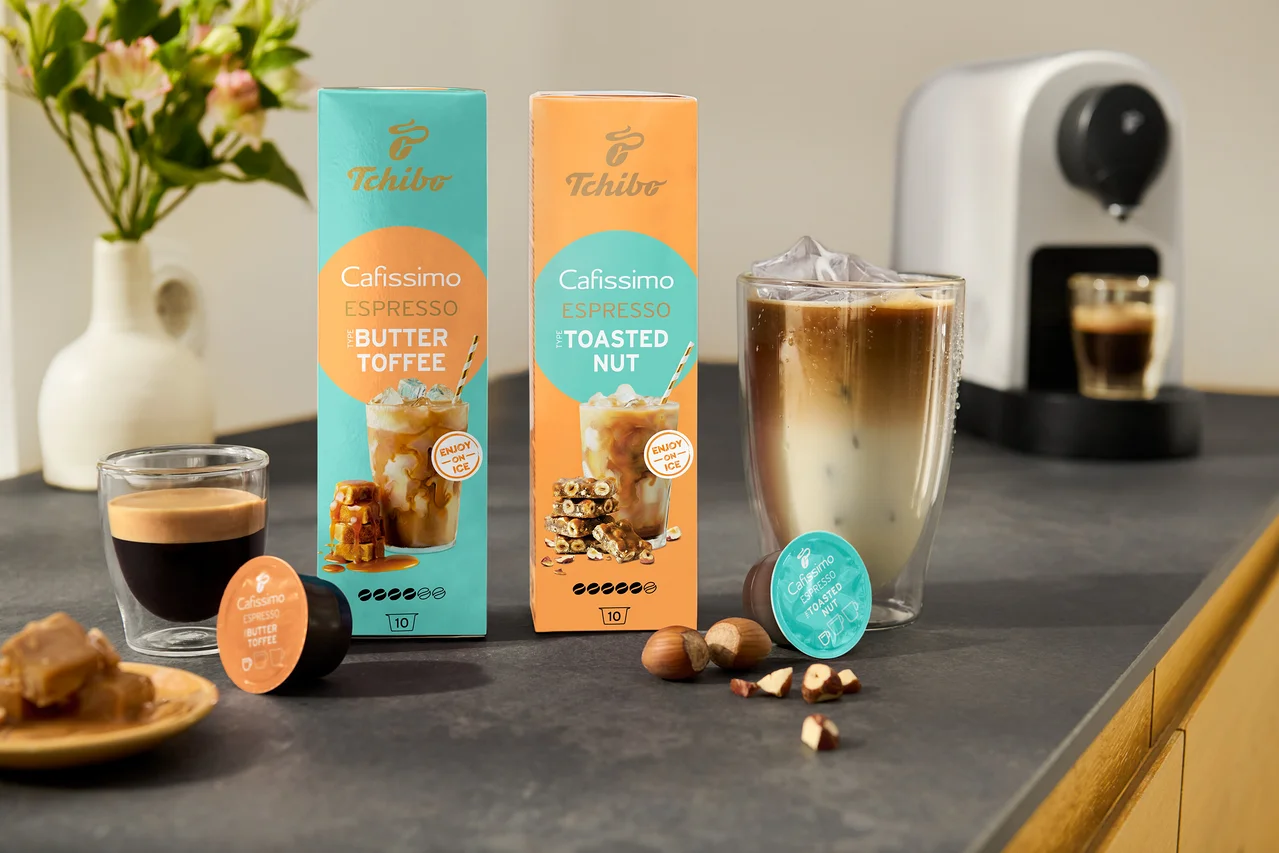 Na kuchynskej doske sú dve balenia Cafissimo Flavoured Espresso, jedno v tyrkysovej farbe s príchuťou Buttertoffee a druhé v oranžovej farbe s príchuťou Toasted Nut, vedľa bieleho kávovaru Cafissimo pure+.