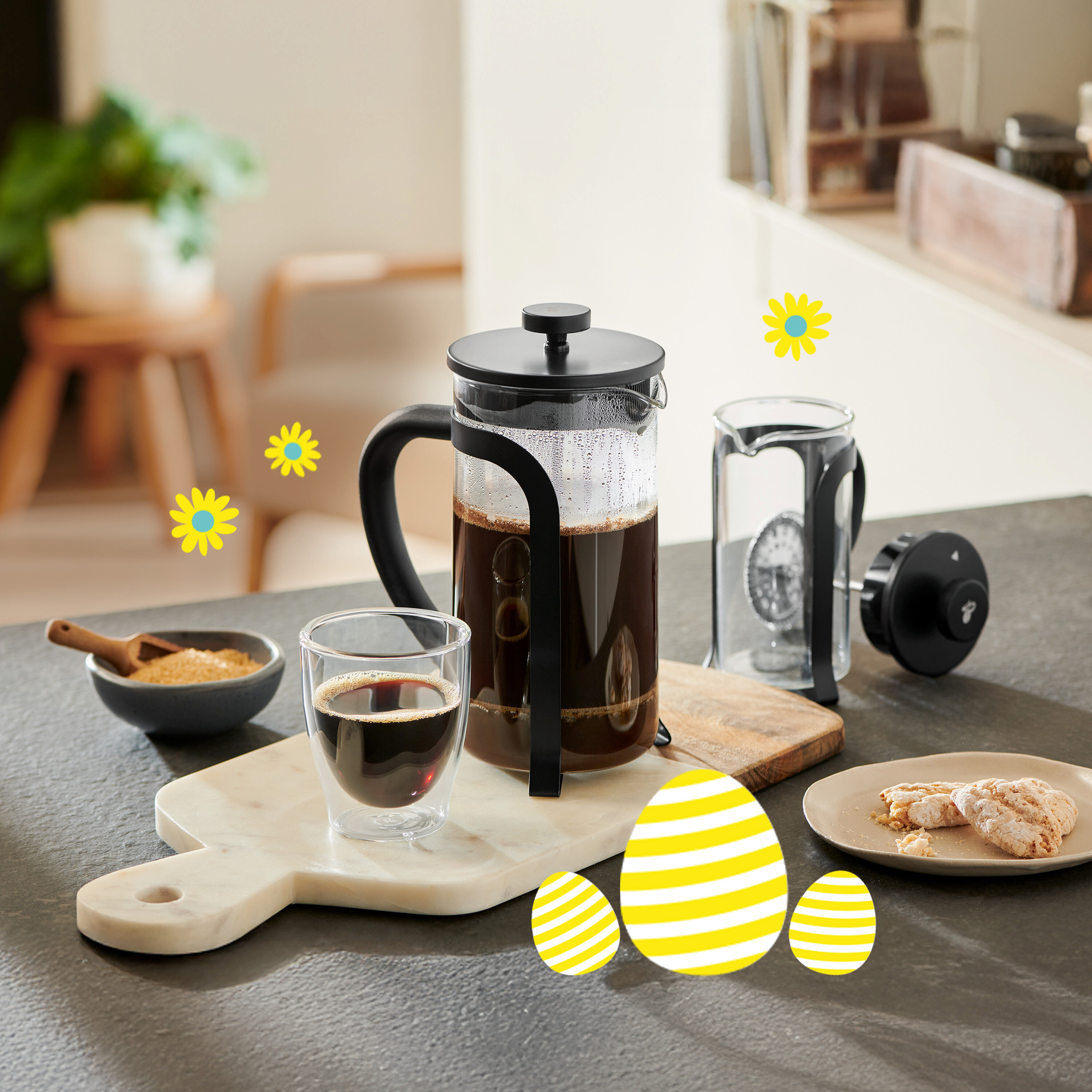 French press vedľa šálky kávy, misky s cukrom a sušienok na stole so žltými veľkonočnými vajíčkami a sedmokráskami.