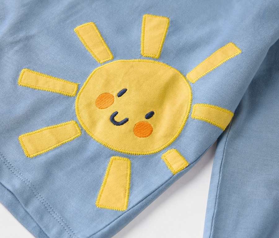 Detailný záber na svetlomodrý odev s našitou žltou slnkom.