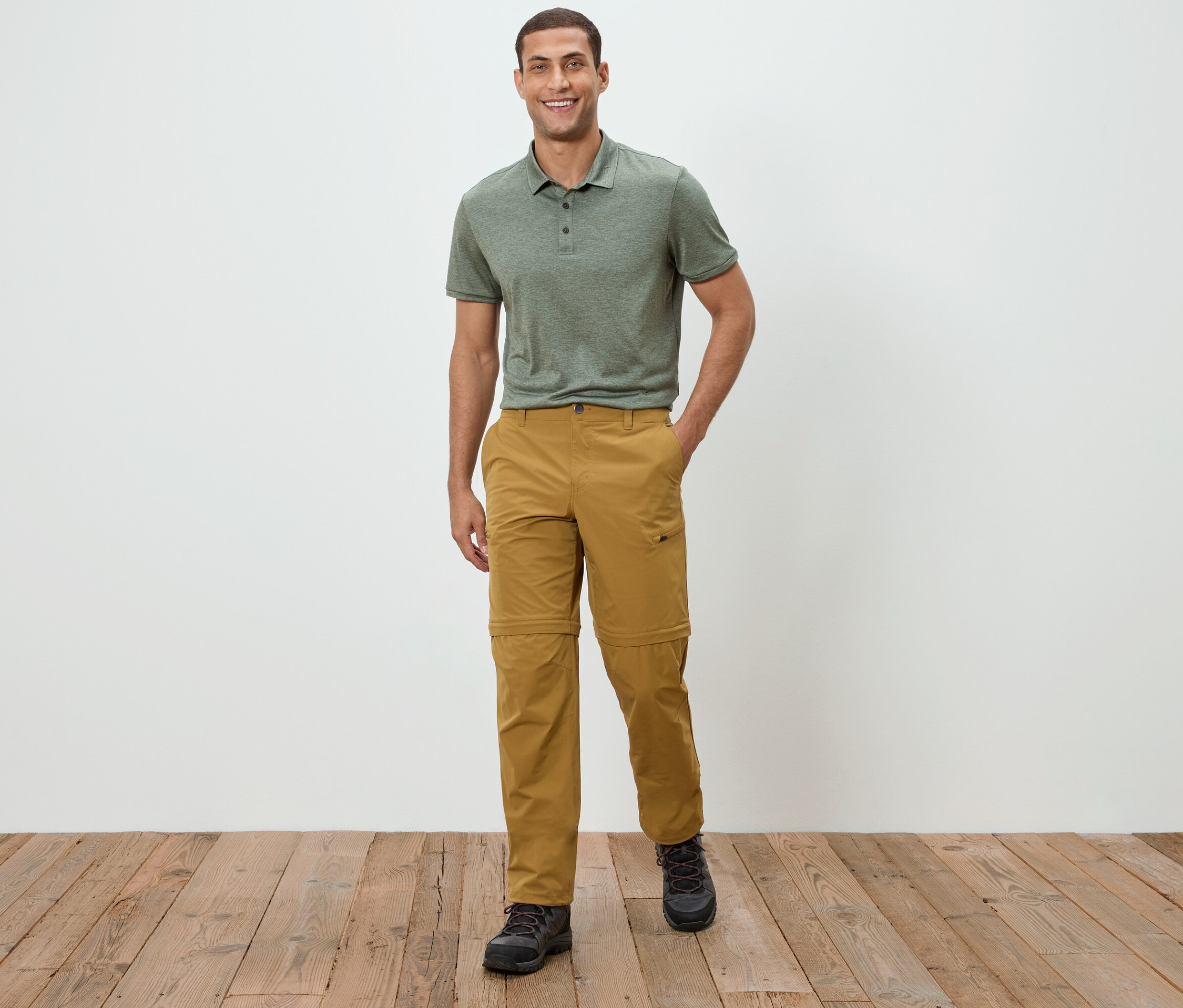 Muž stojí pred bielou stenou a má na sebe zelené polotričko a khaki nohavice.