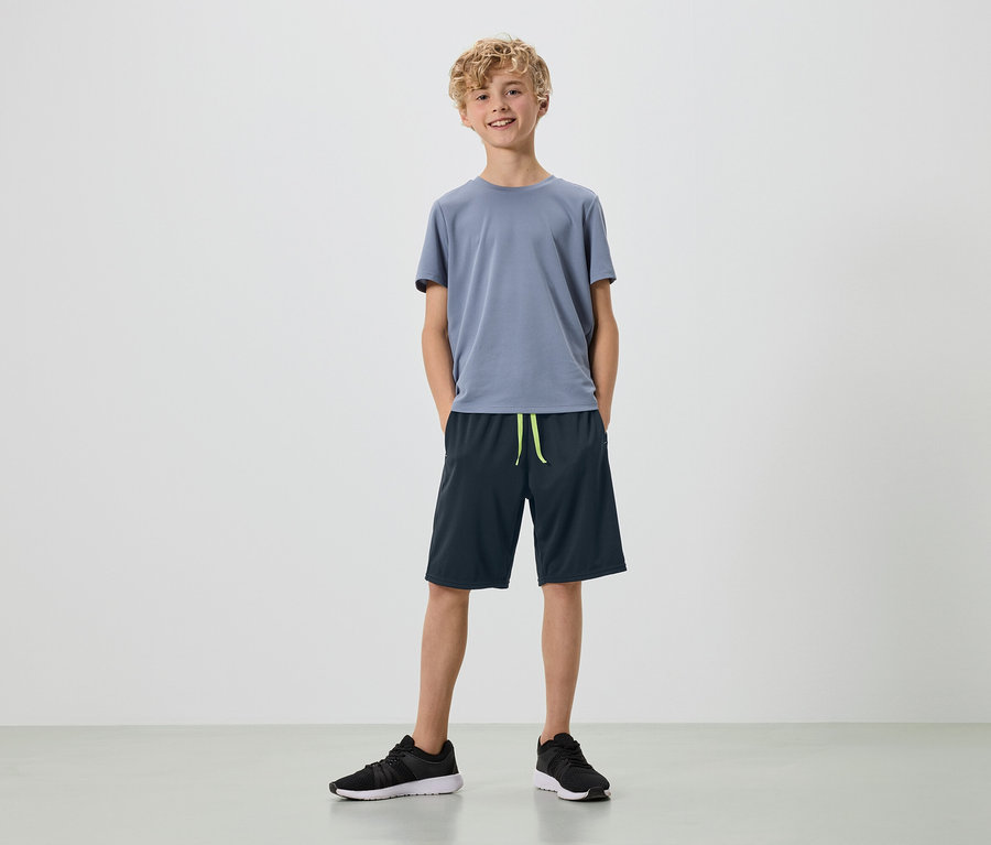 Mladý muž s blond kučerami stojí na bielom pozadí v sivom tričku, tmavomodrých šortkách a čiernych teniskách.