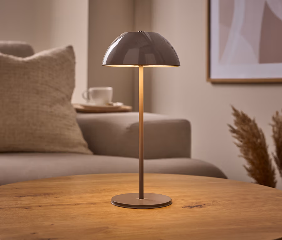 Béžová stolová LED lampa s akumulátorom s kupolovitým tienidlom stojí na okrúhlom drevenom stole. V pozadí je pohovka, šálka a pampová tráva.