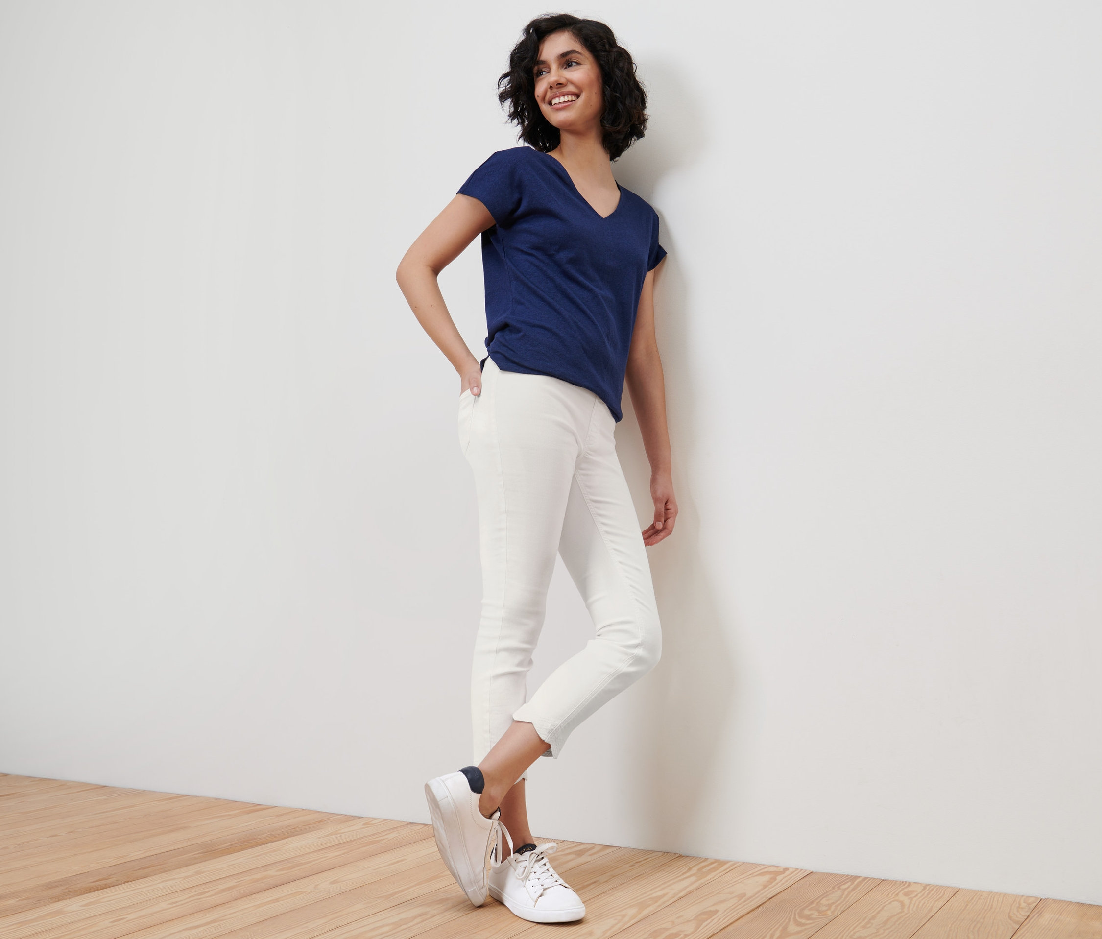 Usmievajúca sa žena opretá o stenu má na sebe modré tričko a biele sedemosminové tregínsy s ažúrovou výšivkou.