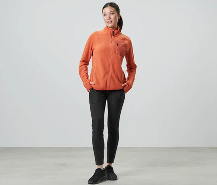 Žena stojí a má ruky vo vreckách outdoorových legín a oranžovej flísovej bundy.