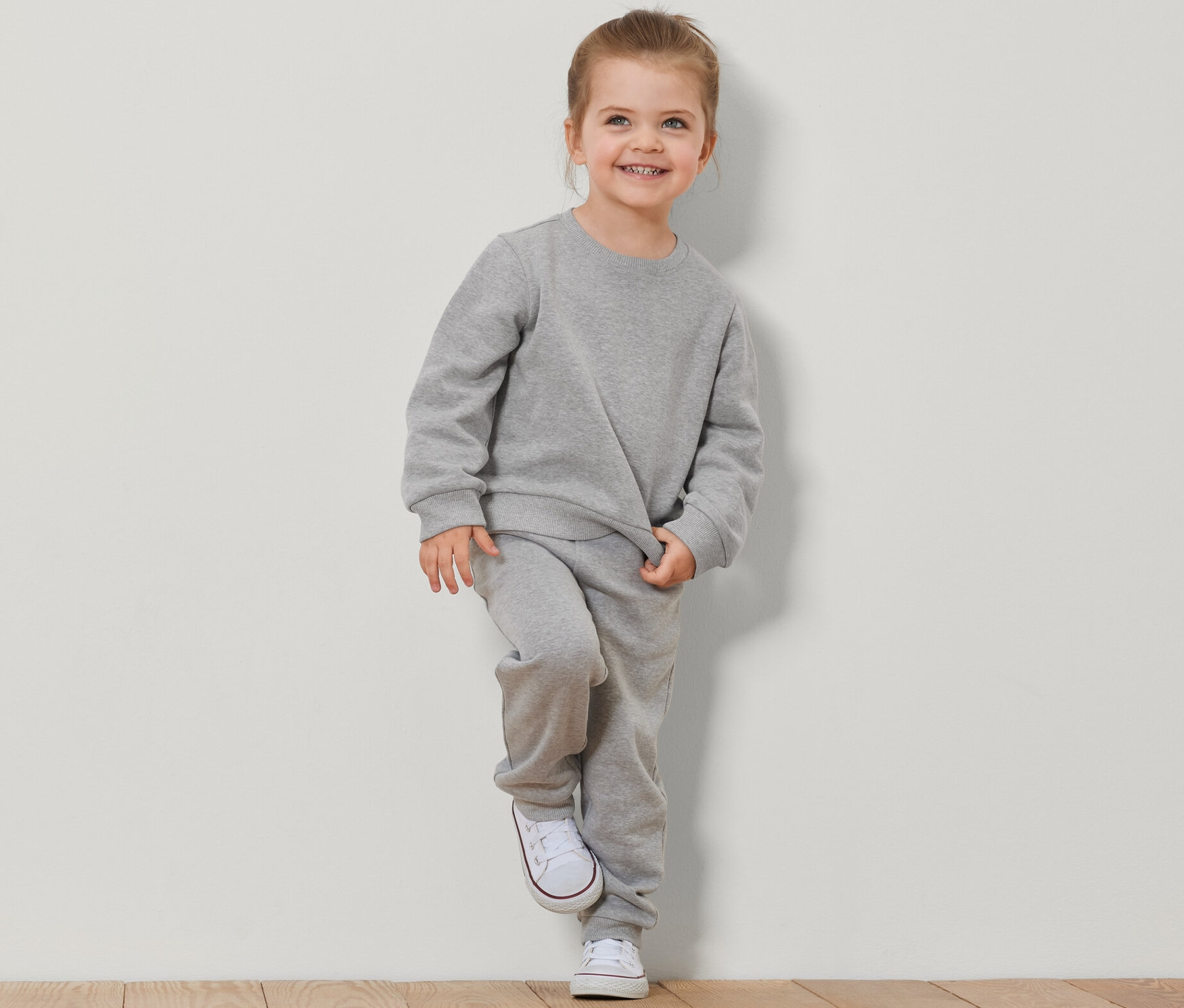 Usmievajúce sa dievča opreté o stenu v súprave Detské mikiny s počesanou vnútornou stranou, 2 ks, šedej farby.