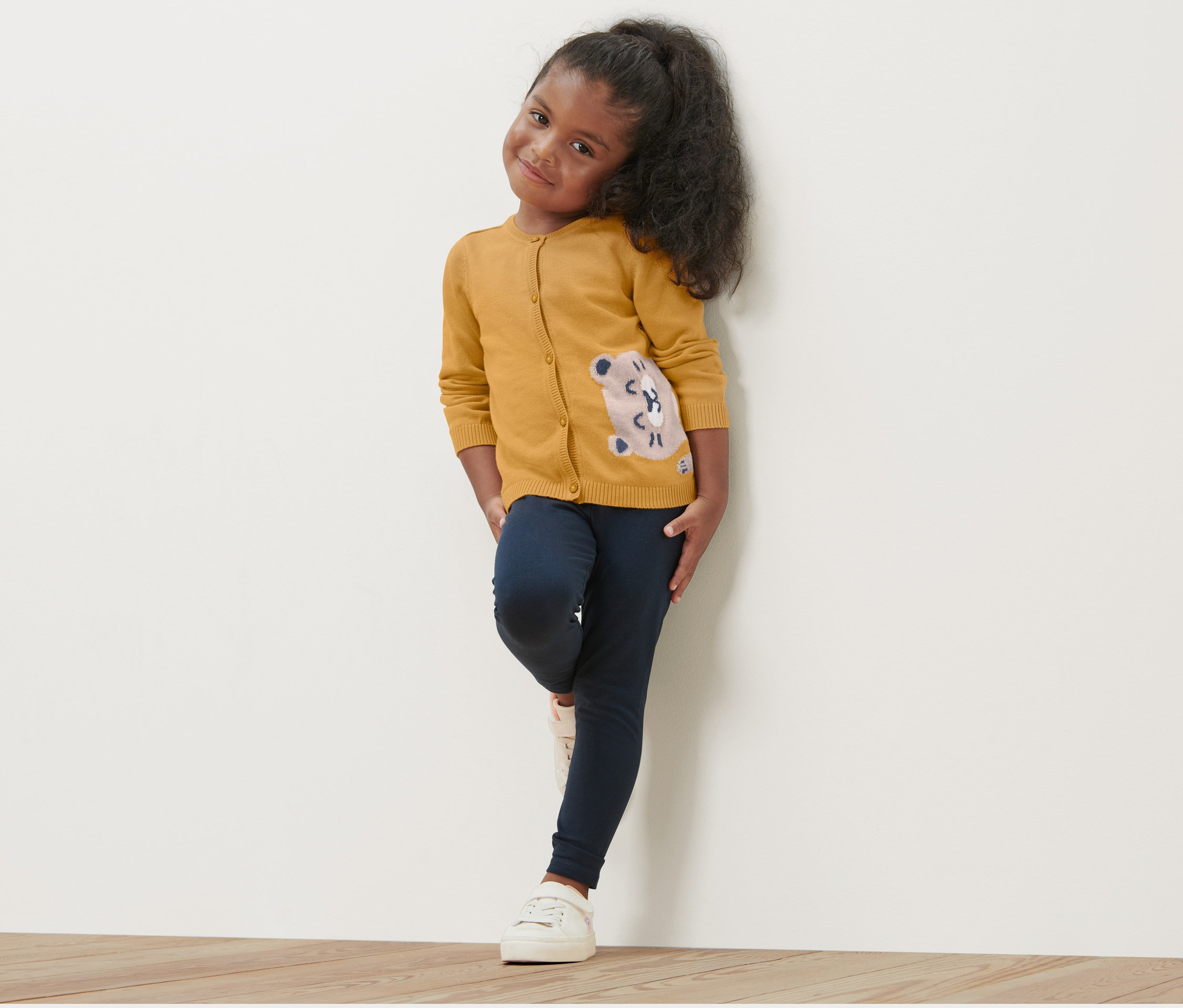 Dievča pózuje v detskom svetriku s motívom vydry, tmavomodrých legínach a bielych teniskách.