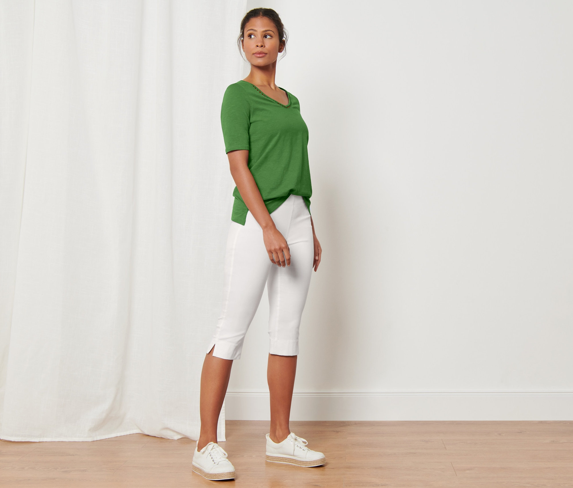 Žena stojaca v miestnosti má na sebe zelené tričko a biele trojštvrťové elastické nohavice.