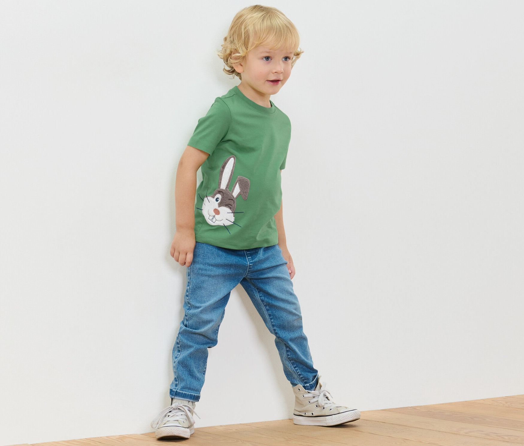 Chlapec stojí pri stene a má na sebe zelené tričko s aplikáciou a rifle.