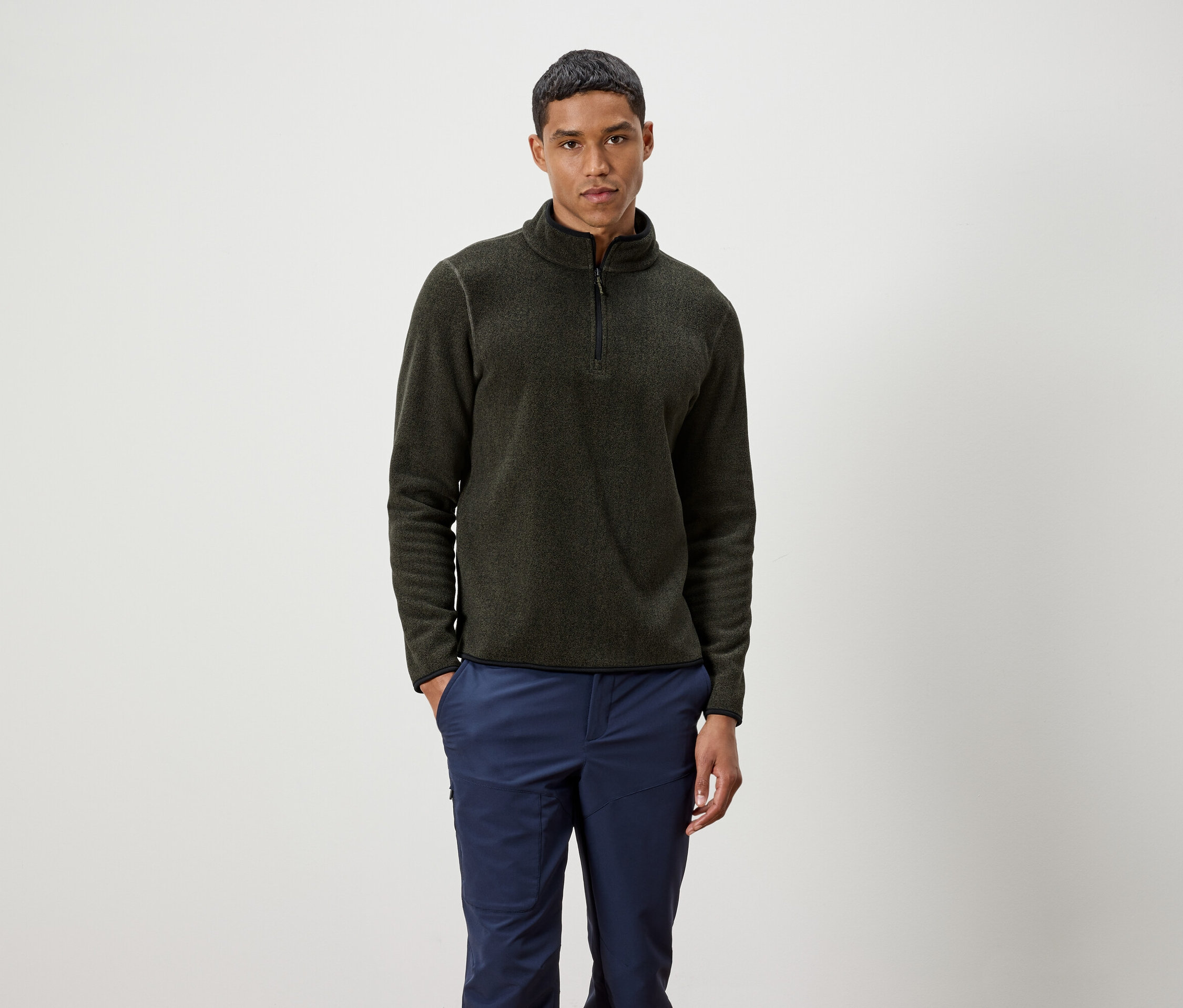 Muž stojí a má na sebe olivový sveter na zips a modré nohavice.