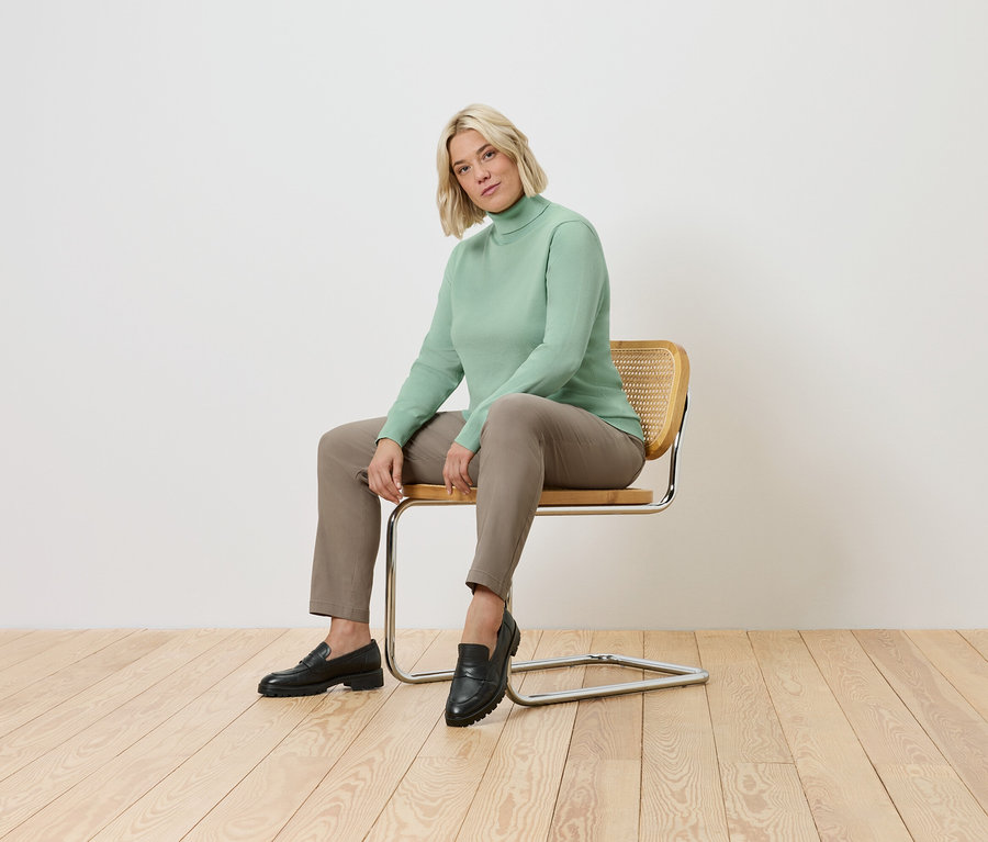 Žena sedí na stoličke. Má na sebe zelený rolák, hnedé nohavice a čierne topánky.