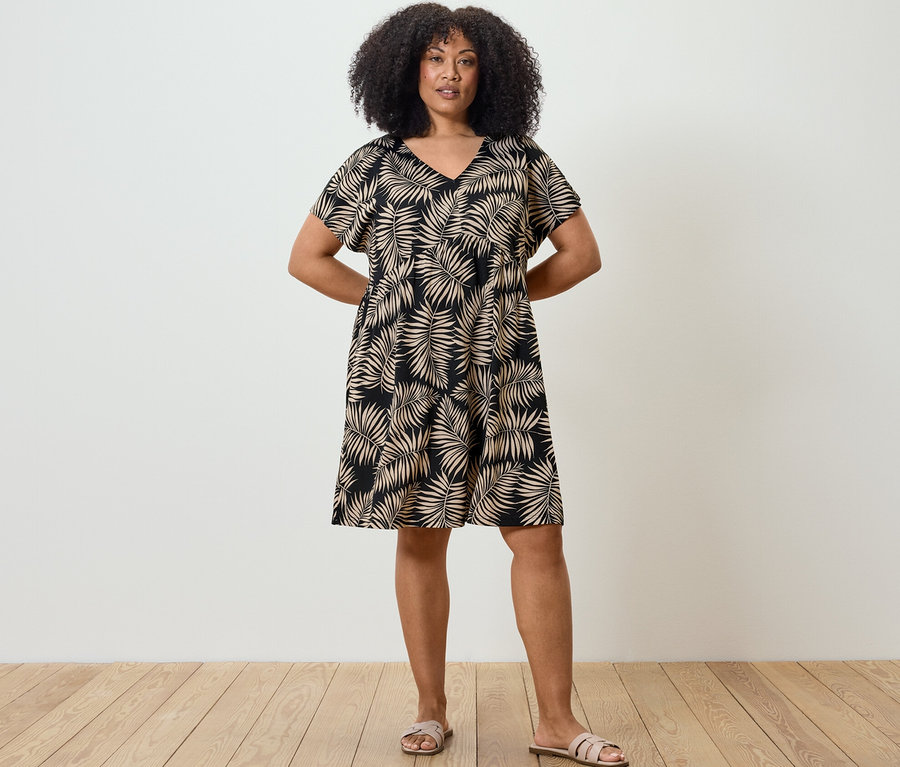 Žena stojí a má ruky za chrbtom. Oblečená je v džersejových šatách s potlačou.