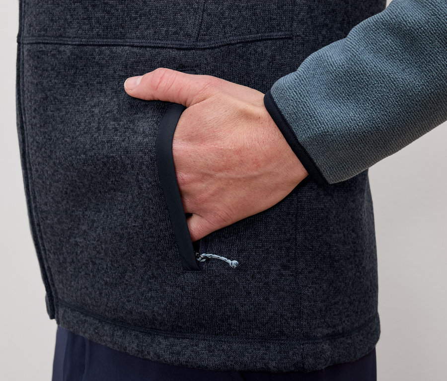 Muž vkladá ruku do vrecka sivo-modrej flísovej bundy.