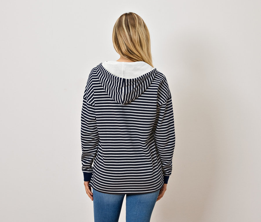 Zozadu je vidieť žena s blond vlasmi, ktorá má na sebe modro-biely pruhovaný pulóver s kapucňou a džínsy.