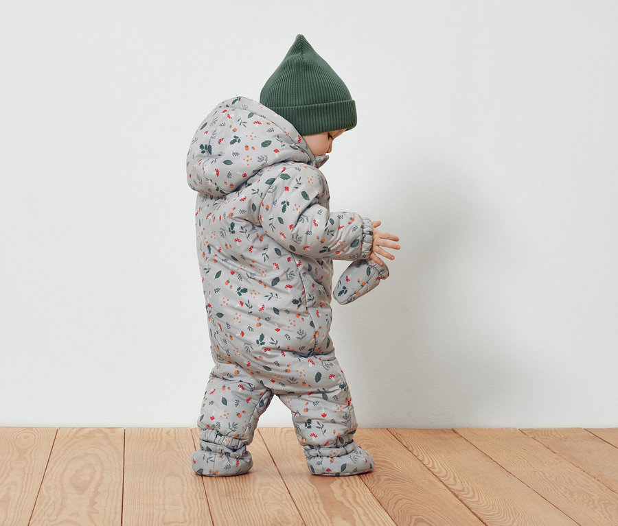 Batoľa v sivom overale s kvetinovým vzorom a zelenej čiapke.