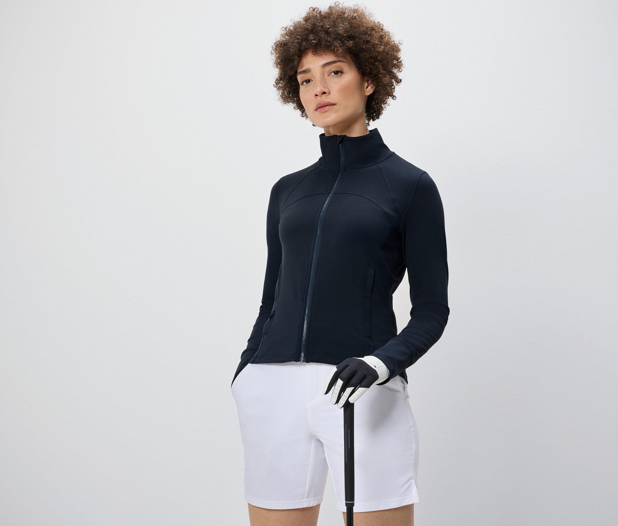 Žena má na sebe funkčnú bundu, biele šortky a drží golfovú palicu.