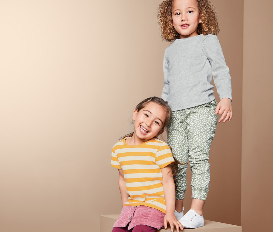 Dve dievčatá pózujú na bloku. Jedna má na sebe sivé rebrované tričko a zelené nohavice. Druhá má žlté pruhované tričko a vínovú sukňu.