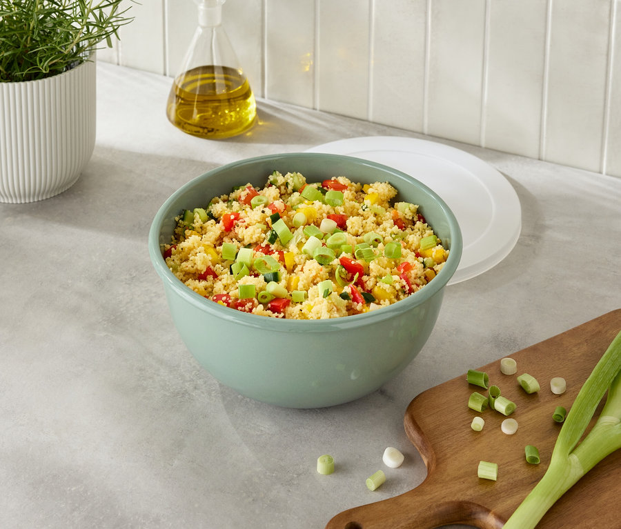 Zelená misa s kuskusom a zeleninou vedľa cibule na doske na krájanie.