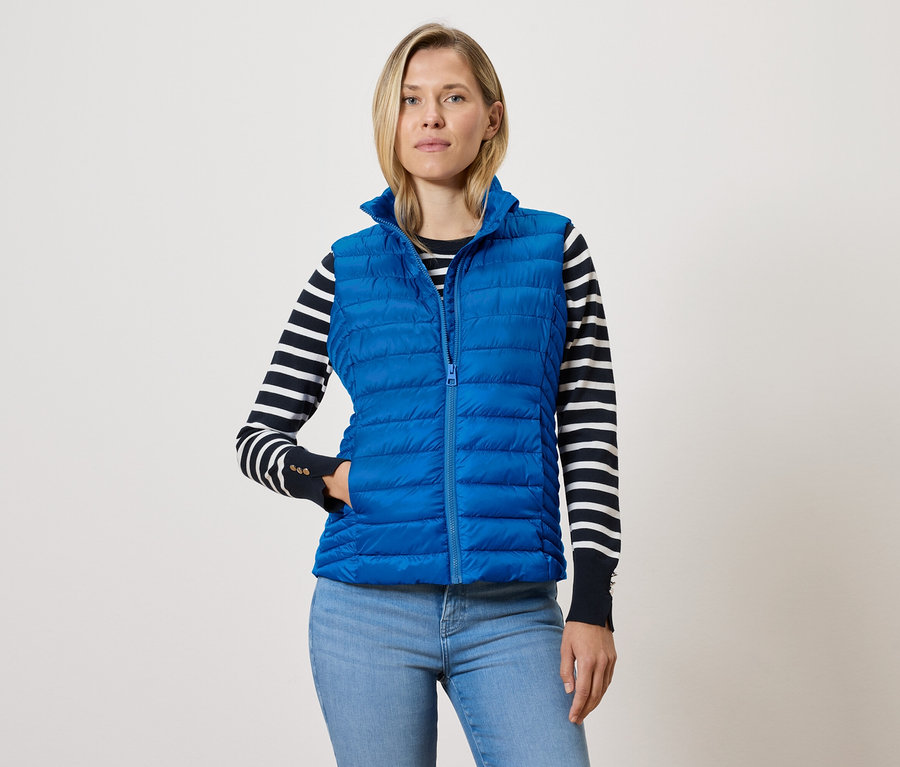 Žena v modrej prešívanej veste so zipsom, svetri s čiernymi a bielymi pruhmi a modrých džínsoch.