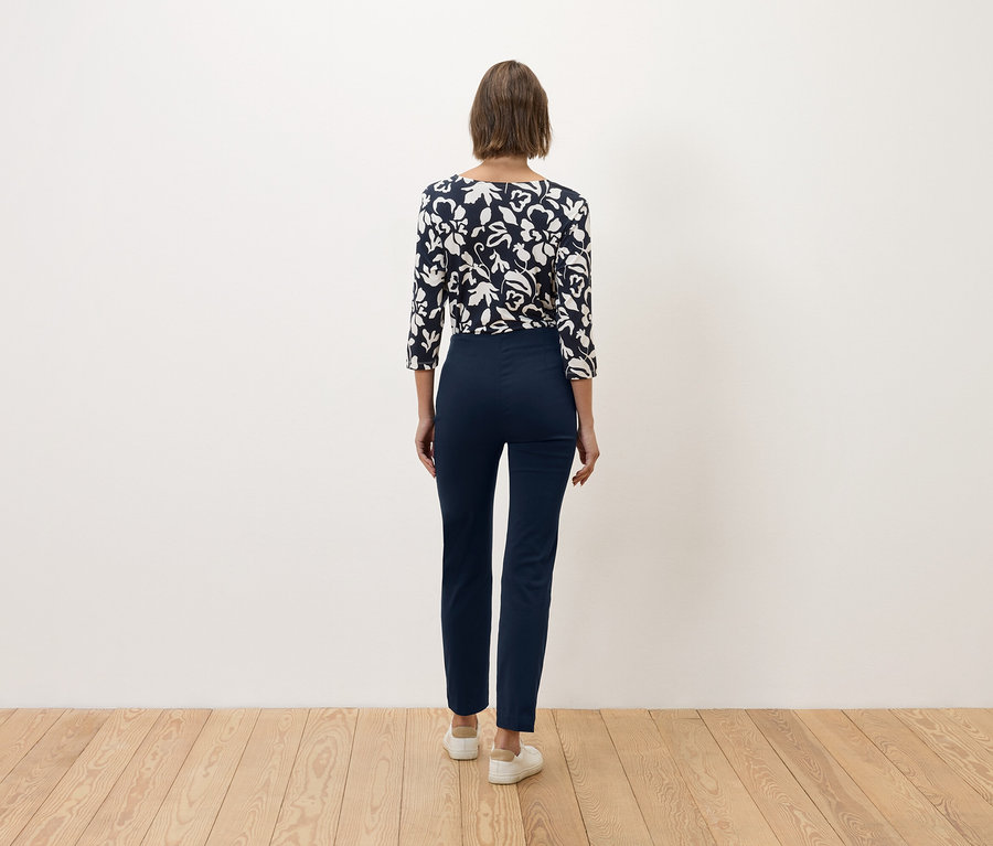 Žena s hnedými vlasmi stojí chrbtom k fotoaparátu a má na sebe tmavomodré nohavice a modro-biely kvetinový top.