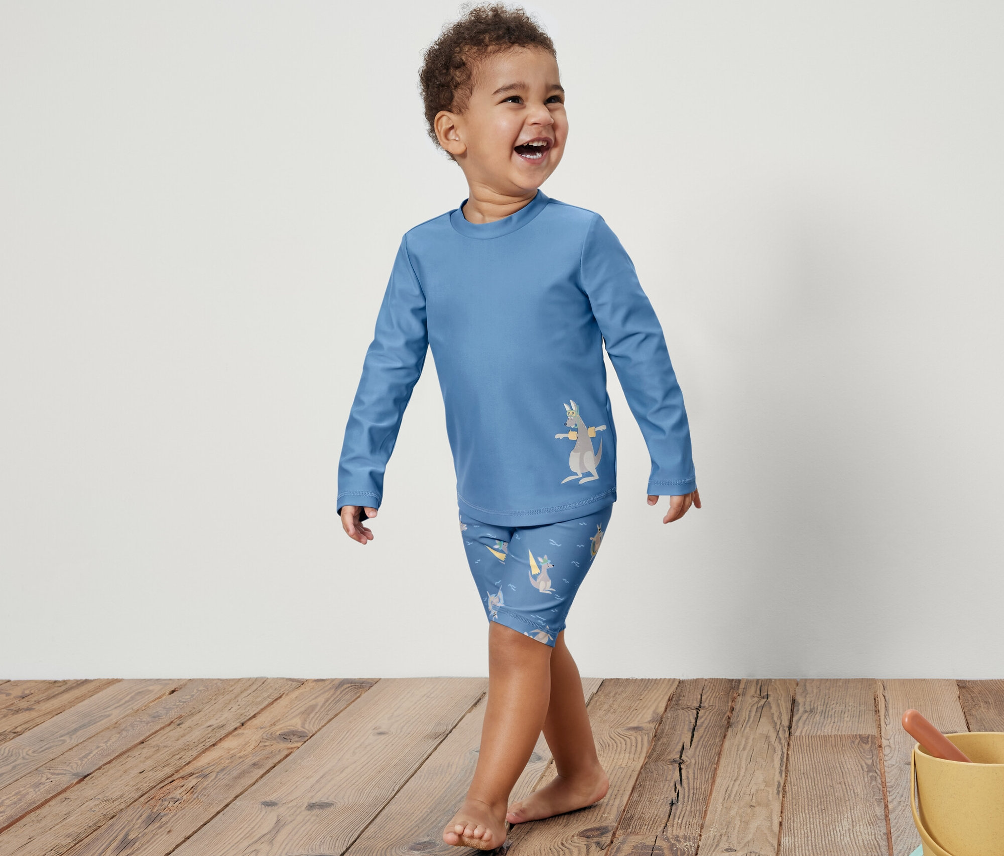 Usmievajúci sa chlapček v modrej detskej súprave s UV faktorom 80 s potlačou kengury kráča po drevenej podlahe. Vpravo je žlté vedierko s hračkami na piesok.