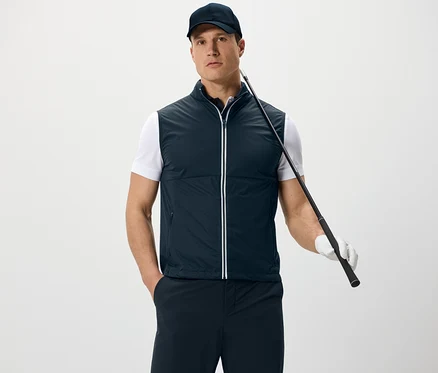Muž má na sebe softshellovú vestu a šiltovku, drží golfovú palicu a stojí pred bielym pozadím.