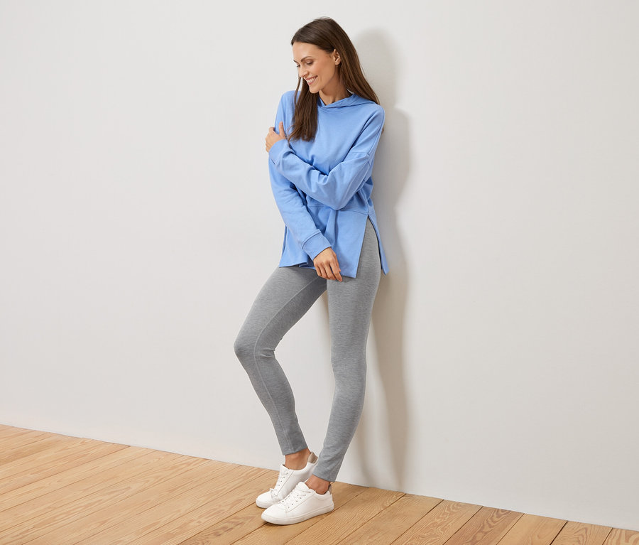 Žena sa opiera o stenu, má na sebe svetlomodrú mikinu s kapucňou s bočnými rozparkami, sivé legíny a biele tenisky.