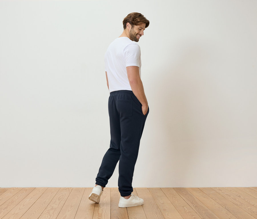 Muž v bielom tričku a modrých teplákoch stojí na drevenej podlahe.