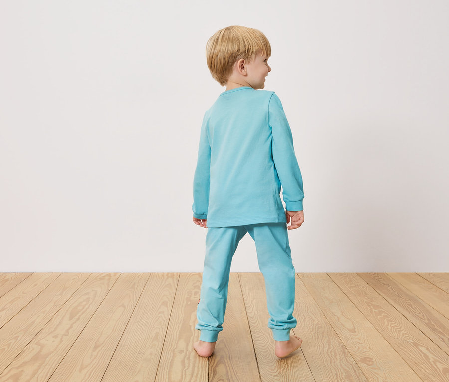 Malý chlapec zozadu, oblečený v svetlomodrom pyžame, stojí na drevenej podlahe pred bielou stenou.