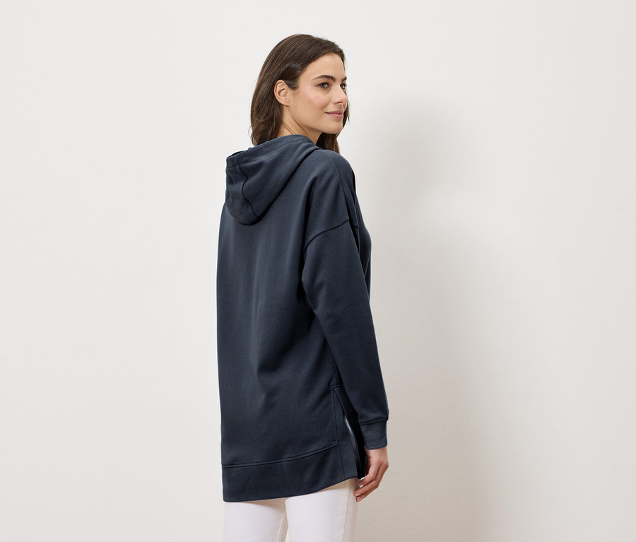 Žena nosí dlhú tmavomodrú mikinu s kapucňou a biele nohavice, pohľad zboku.