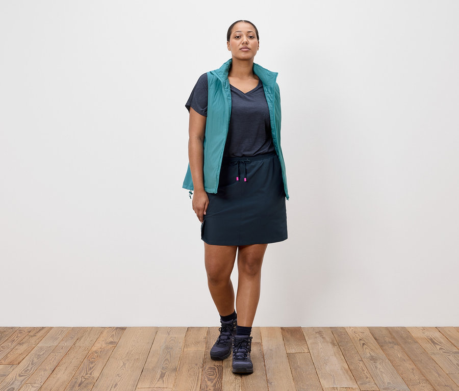 Žena stojí pred bielou stenou, má na sebe tmavomodrú sukňu, tmavomodré tričko, petrolejovú vestu a tmavomodré turistické topánky.