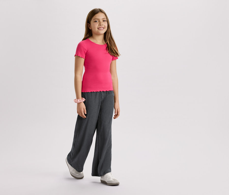 Dievča má na sebe ružové tričko, sivé nohavice a tenisky na bielom pozadí.