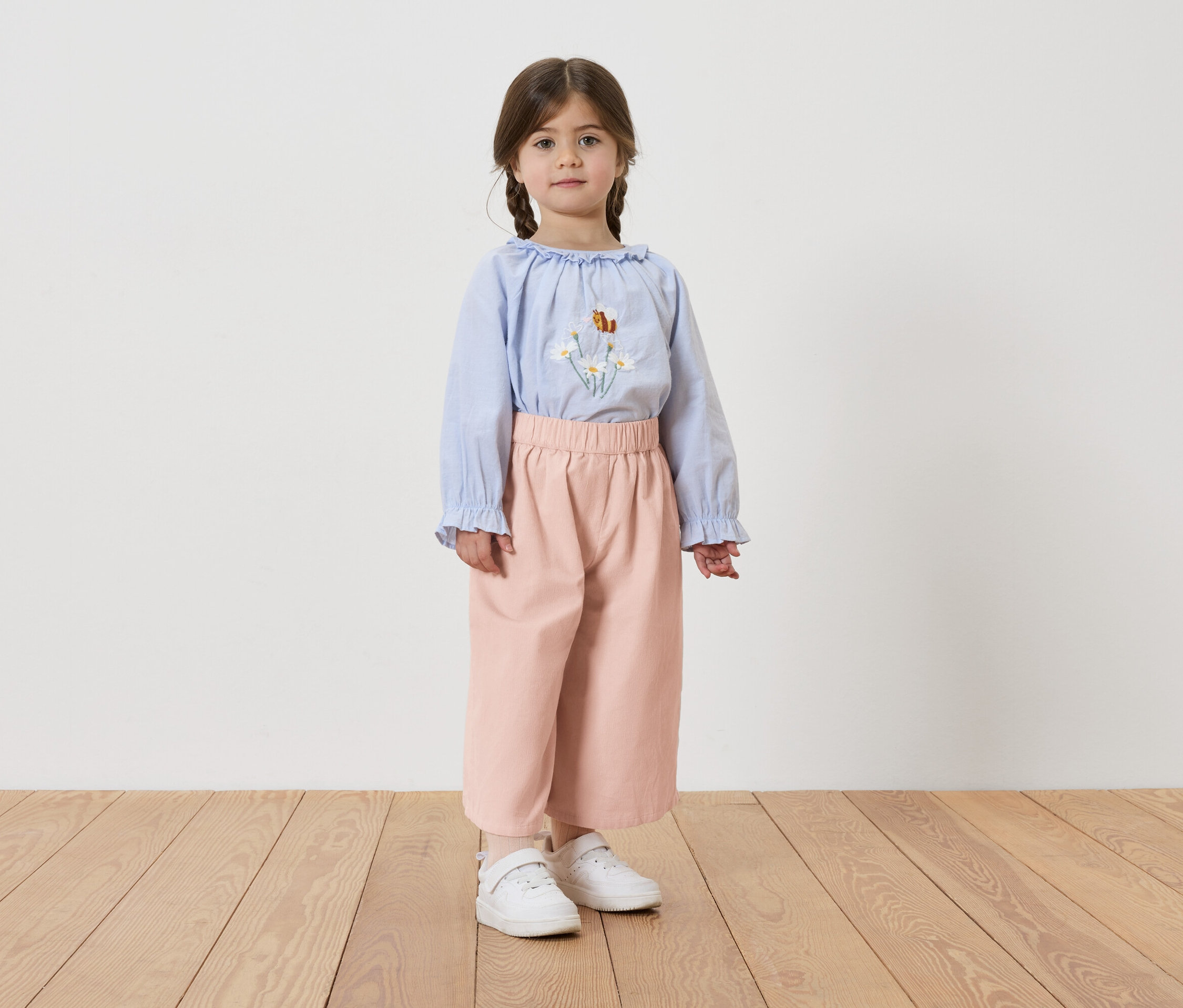 Dievča s vrkočmi má na sebe svetlomodrú blúzku s kvetinovou výšivkou, ružové nohavice a biele tenisky.