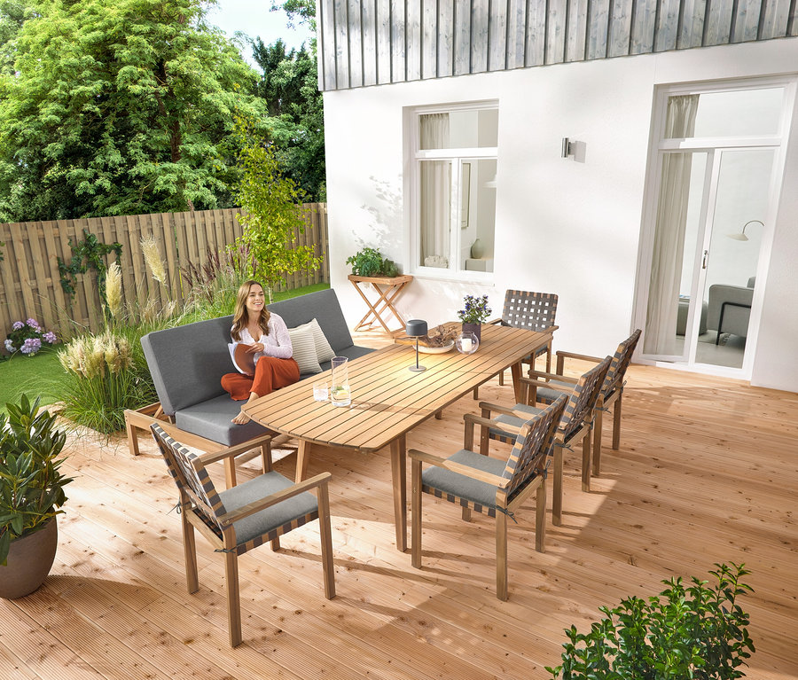 Žena sedí na záhradnej lavici 2 v 1 ‒ dvojitom ležadle. Na rozkladacom záhradnom stole so zaoblenými čelami, cca 180‒240 cm, stojí sivá solárna stolná lampa. K dispozícii je aj záhradný stolík a stohovateľné záhradné stoličky s pásovým výpletom.