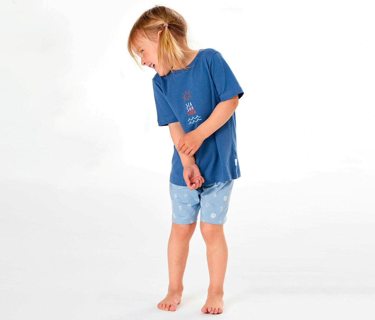 Dievča nosí krátke detské pyžamo SCHIESSER s modrým tričkom a vzorovanými šortkami, usmieva sa nabok.