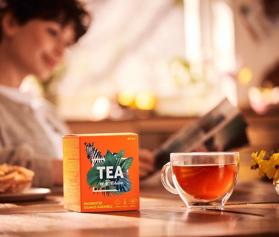 Žena číta noviny pri stole s šálkou čaju a balením čaju TEA by Tchibo Rooibos s príchuťou pomaranča a karamelu.