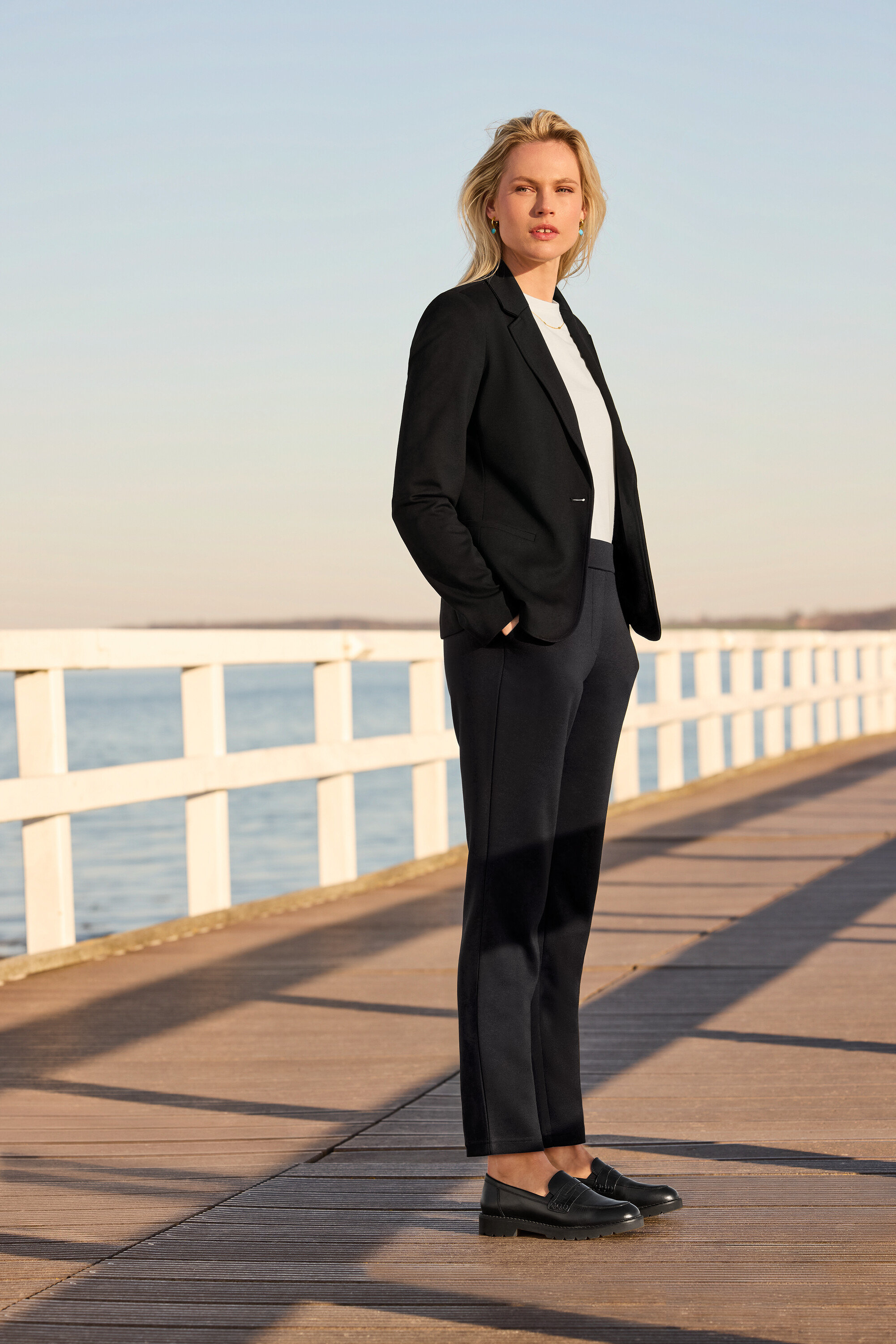 Žena stojaca na móle má na sebe tepláky, jednoduché biele tričko, džersejové sako a kožené mokasíny. Má kreoly s príveskami z kamienkov a retiazku s navetou.