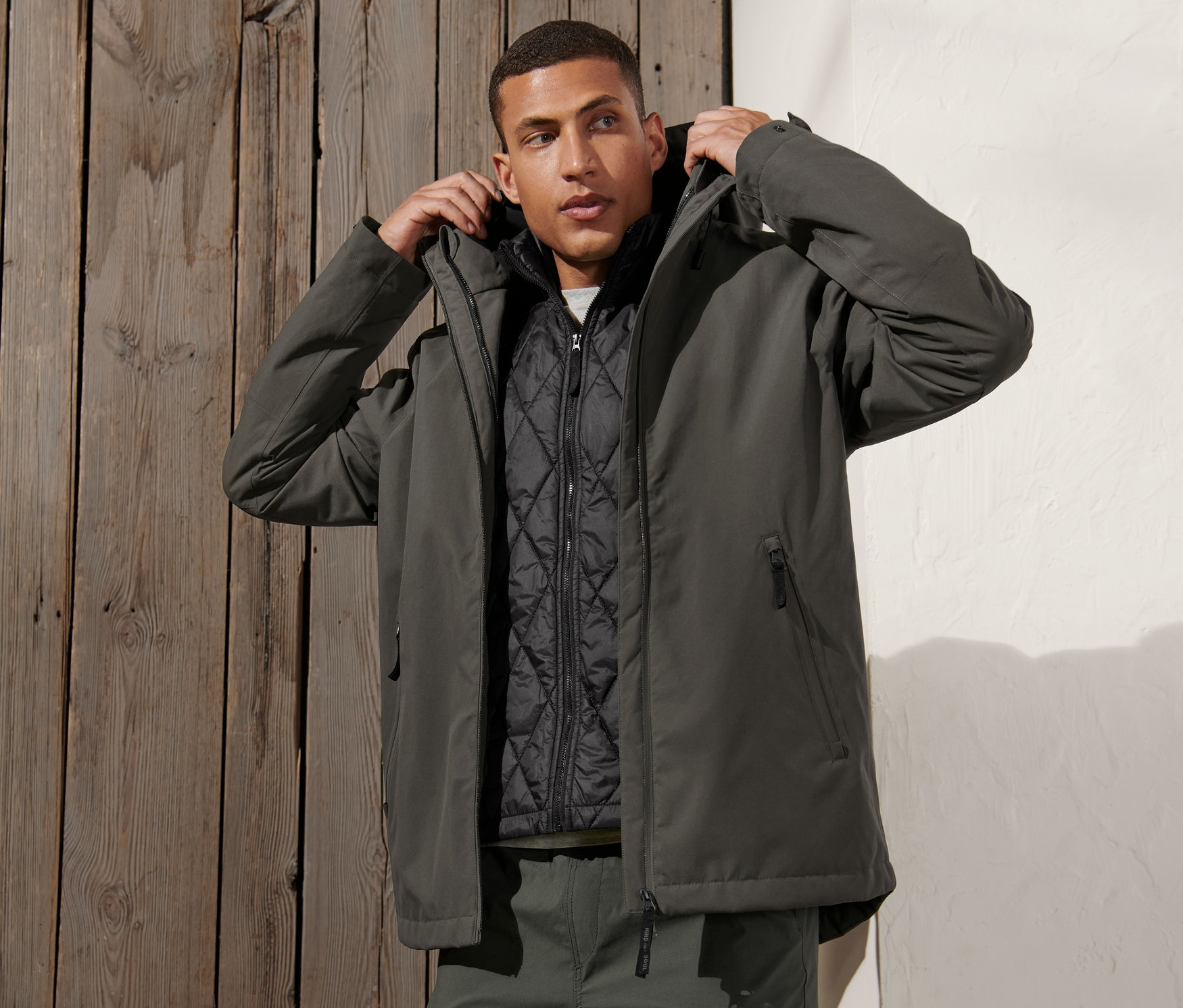 Muž si upravuje kapucňu šedozelenej outdoorovej bundy 3 v 1 s vestou.