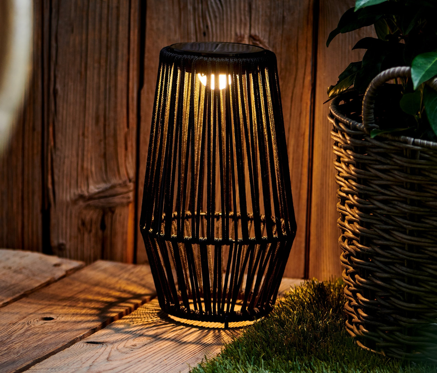 Čierna solárna lampa stojí na drevenej palube vedľa prúteného koša s rastlinou.