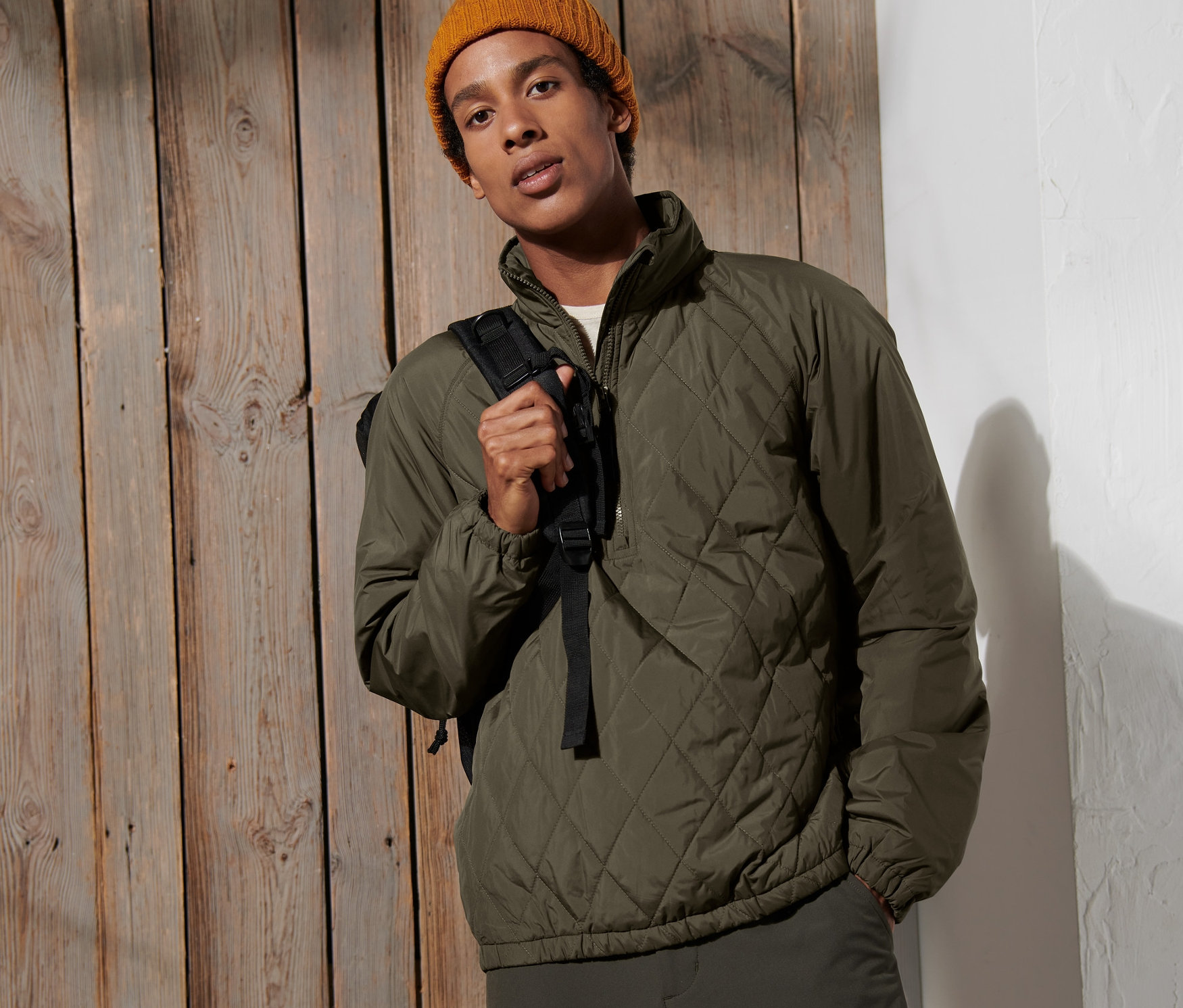 Muž pózuje na pozadí drevenej steny a má na sebe vetruvzdornú bundu a žltú čiapku.