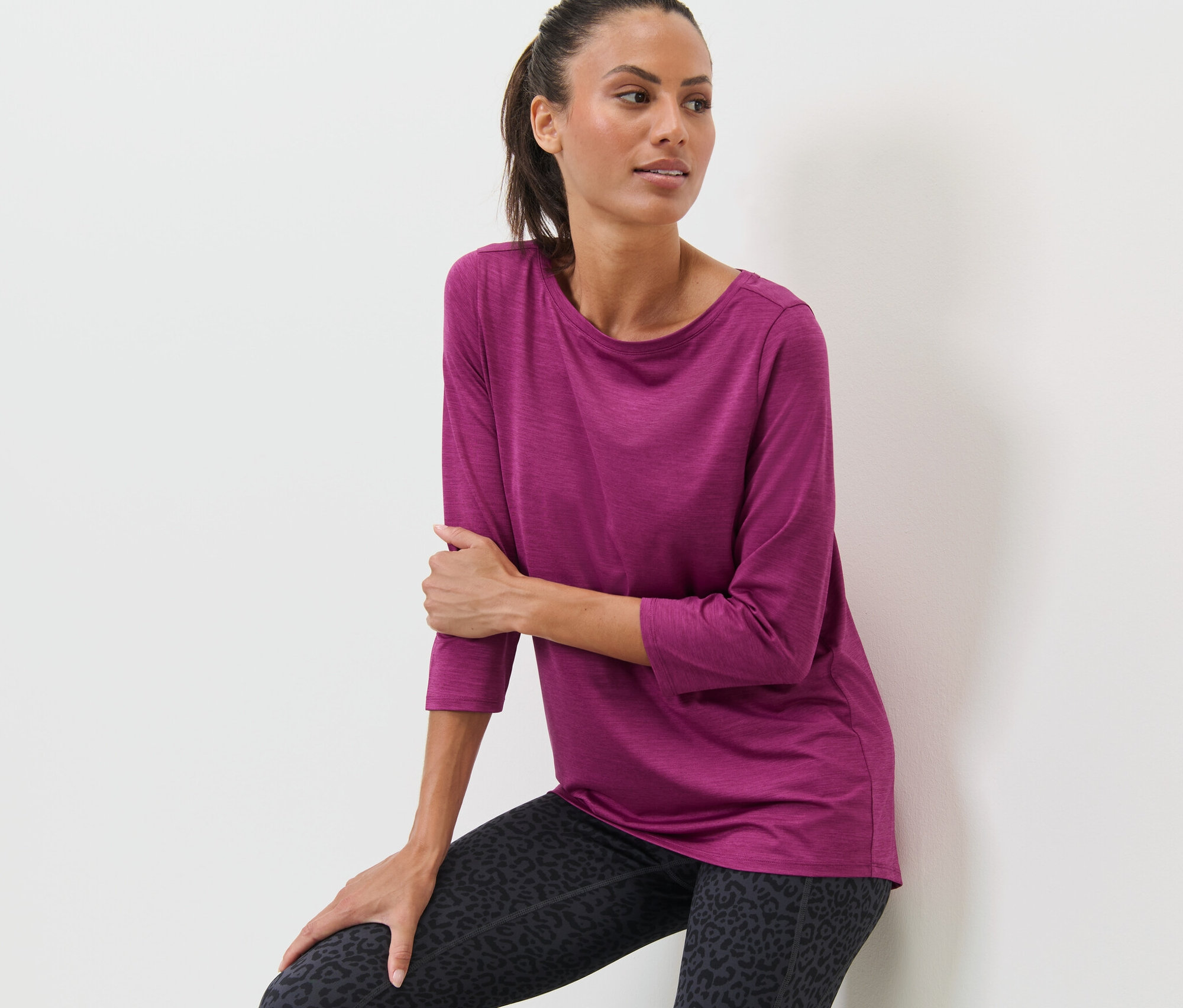 Žena opierajúca sa o stenu má na sebe ružové funkčné tričko s trojštvrťovými rukávmi a legíny so zvieracou potlačou.